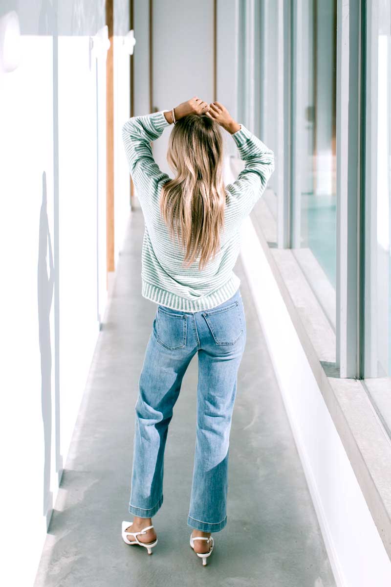 Woman in a Billie Stitch Stripe Knit in sage and blue jeans standing back on in a modern indoor setting.