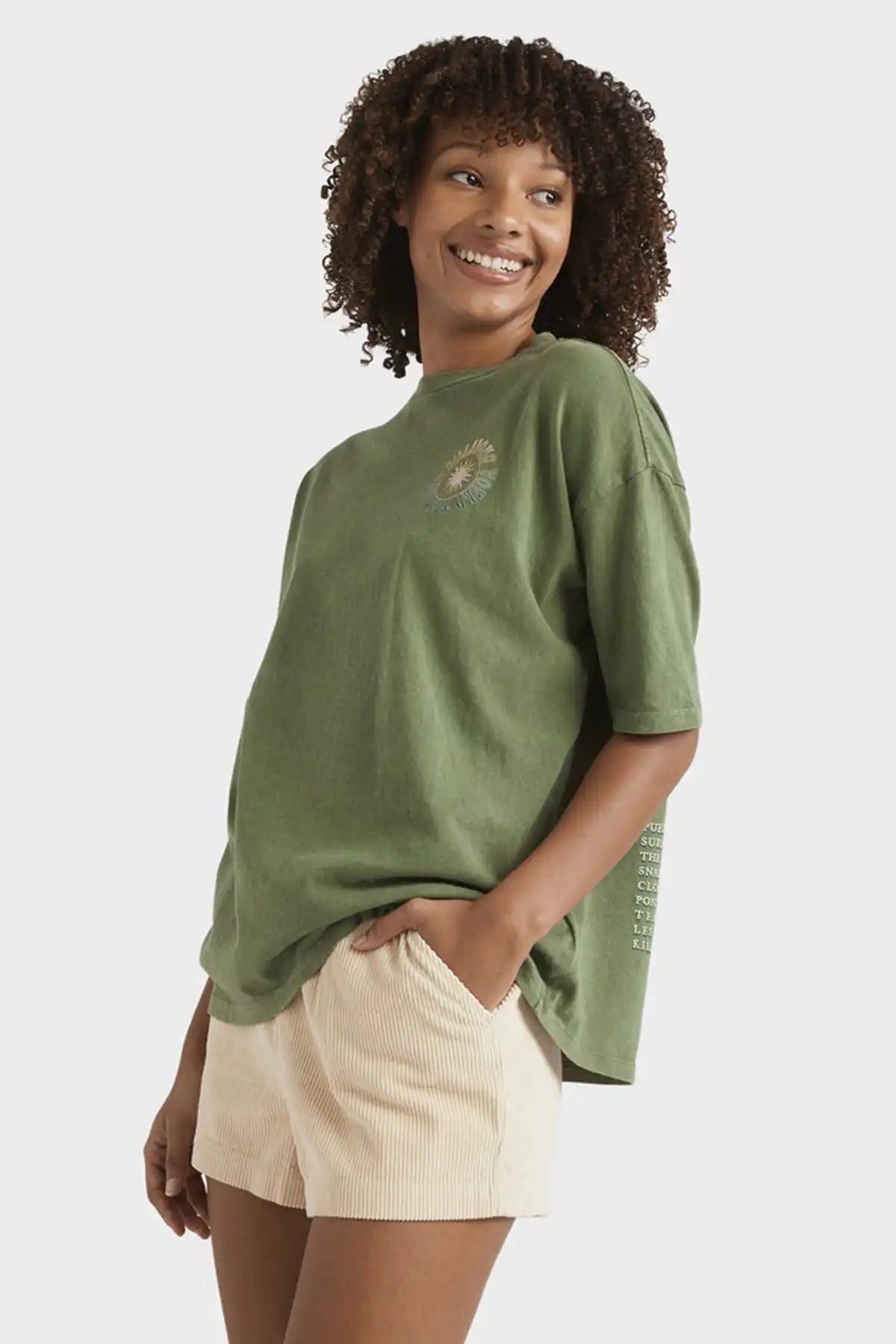 Woman wearing a Billabong Women's Take A Tour oversized t-shirt and beige shorts on a white background