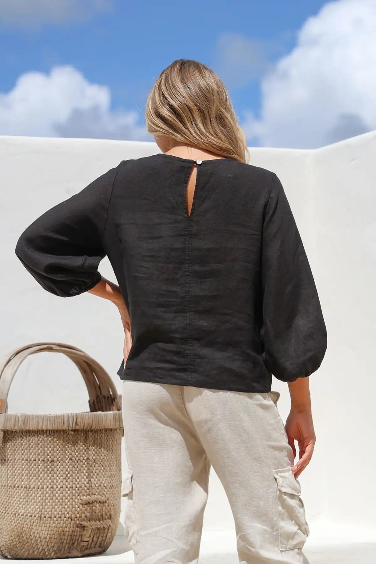 Person wearing a Giovanna Italian Linen Top in black and beige pants standing against a white wall with a blue sky back view.