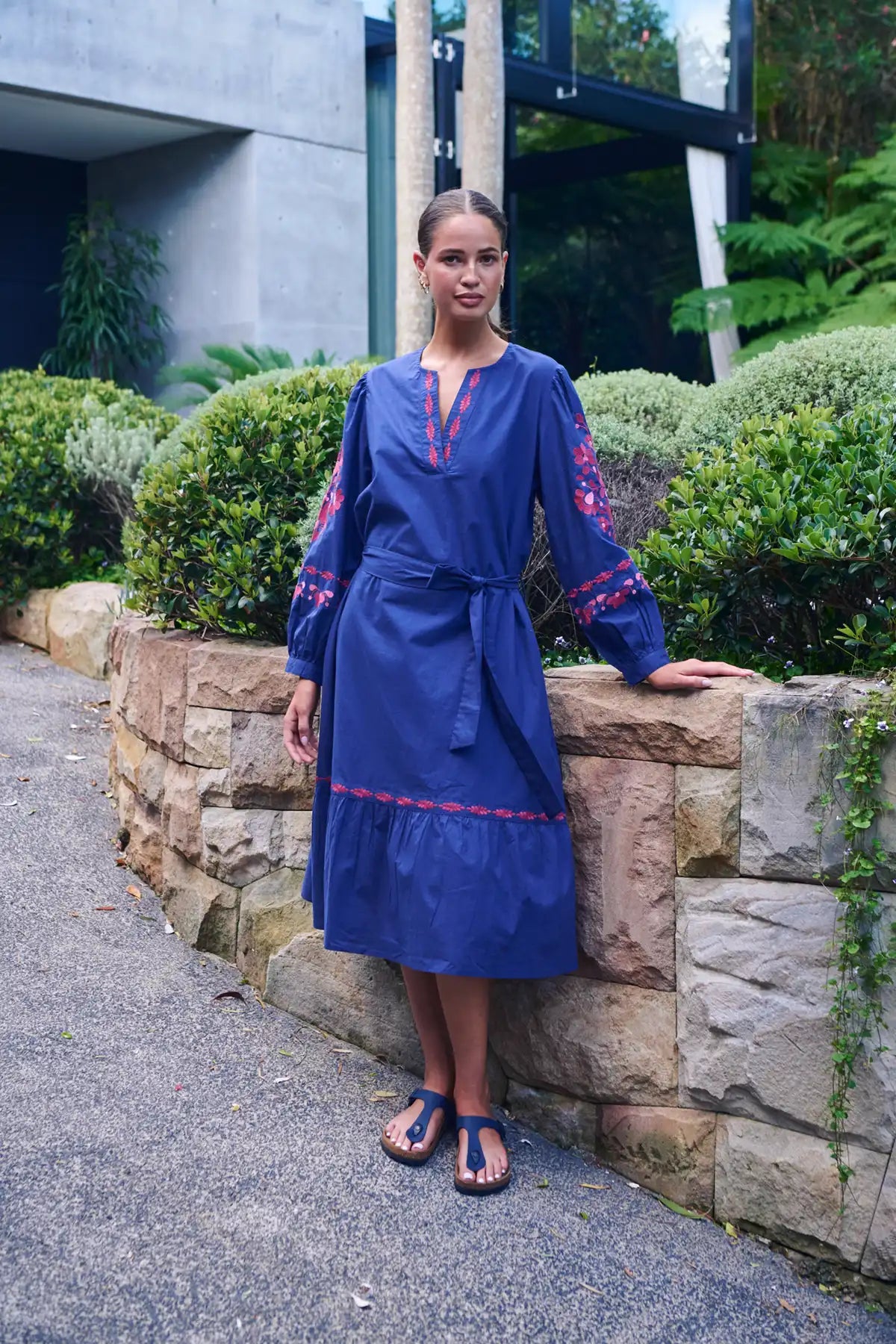 woman wearing a Ink Rose Embroidered Midi Dress leaning against a rock wall in the garden