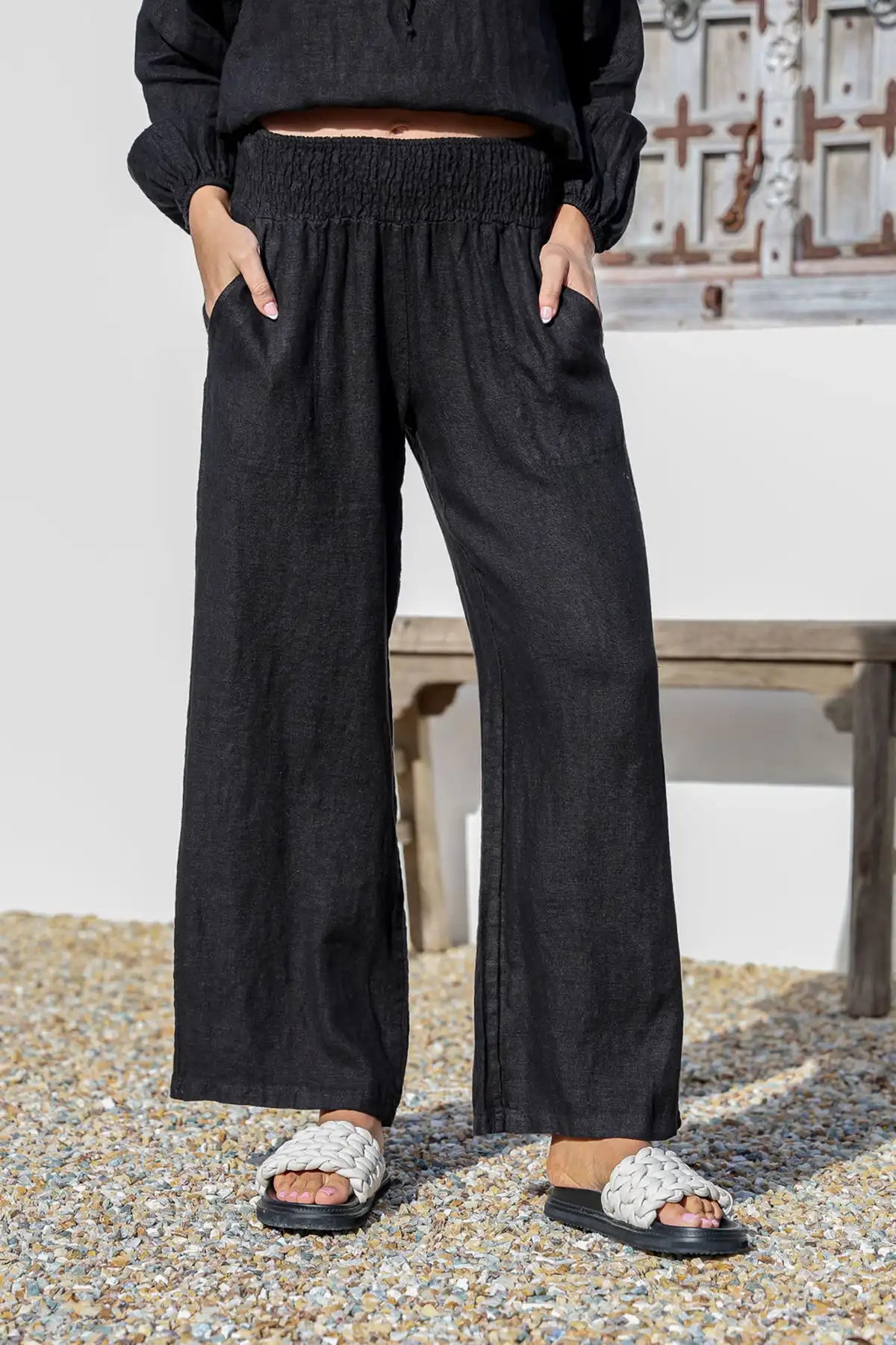 Person wearing Isabella Italian Linen Pant in Black, wide-leg pants standing on a pebbled surface with a wooden bench in the background, front detailed view.
