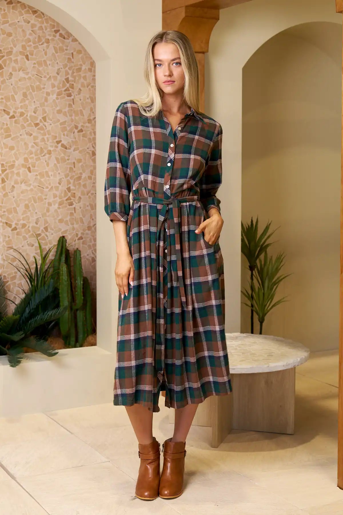 Woman wearing Laura Midid Dress in Forest Plaid standing front on in an indoor setting with plants and a bench.