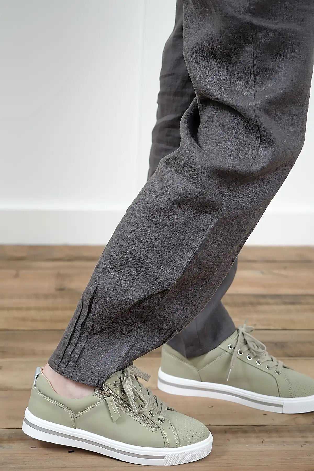 Person wearing Lina Linen Pull-On Pants in Charcoal and green sneakers on a wooden floor, detail view of the pleated cuffs.