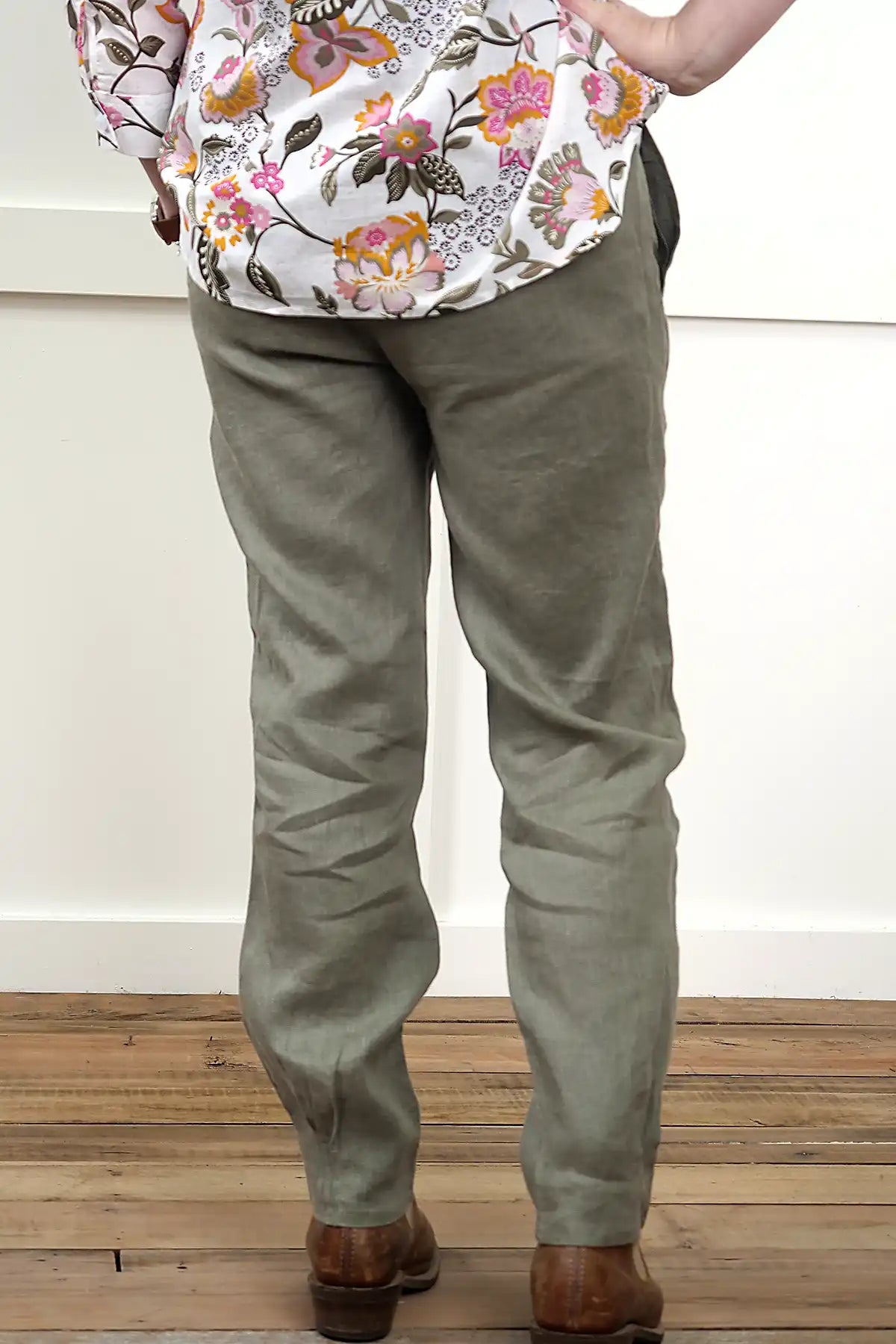 Person wearing a floral top and Lina Linen Pull-On Pants in Olive from behind on a wooden floor with a white wall background
