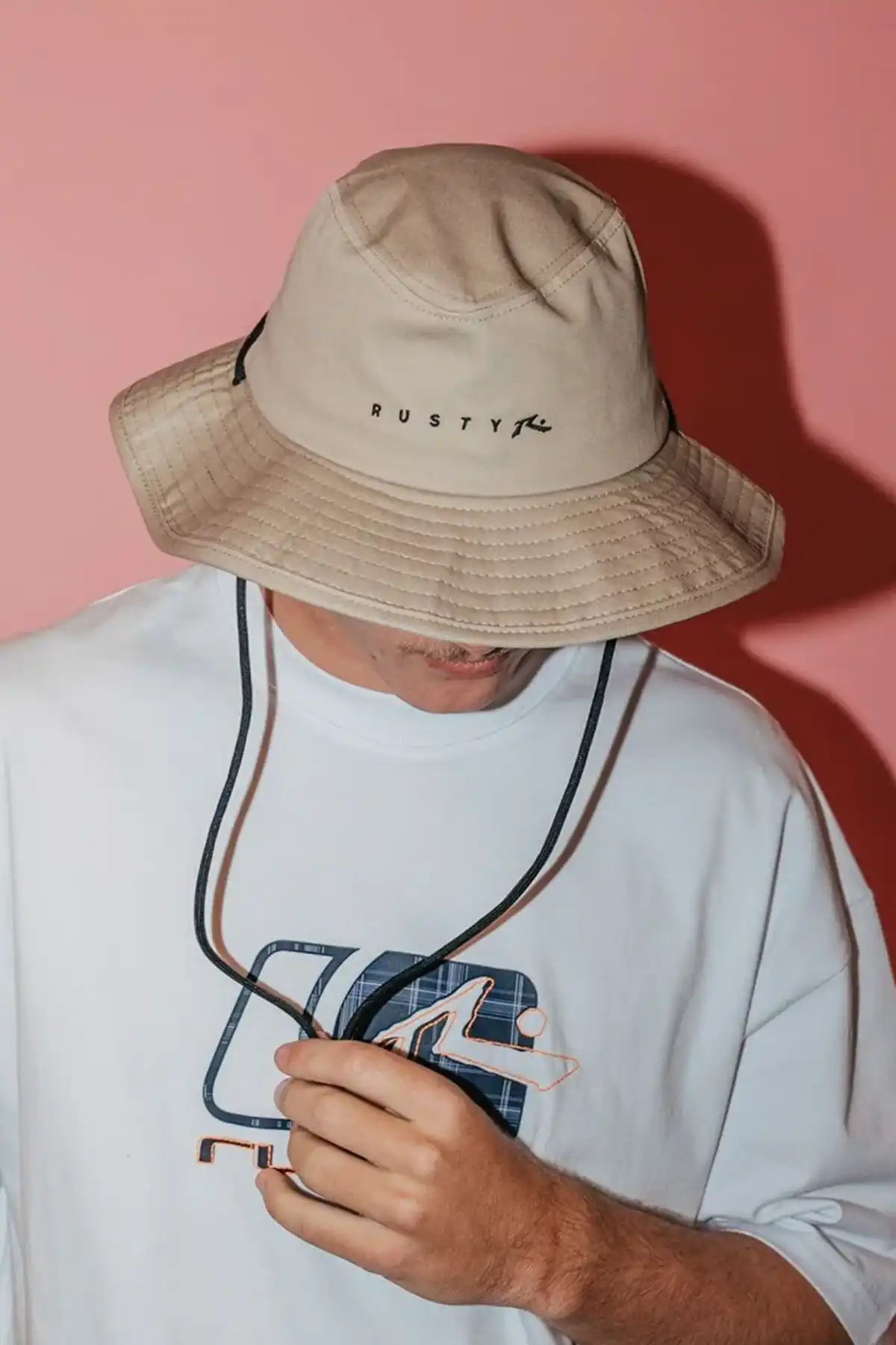 Person wearing a Rusty Bradman Wide Brim Hat in Abbey Stone with 'Rusty' branding against a pink background