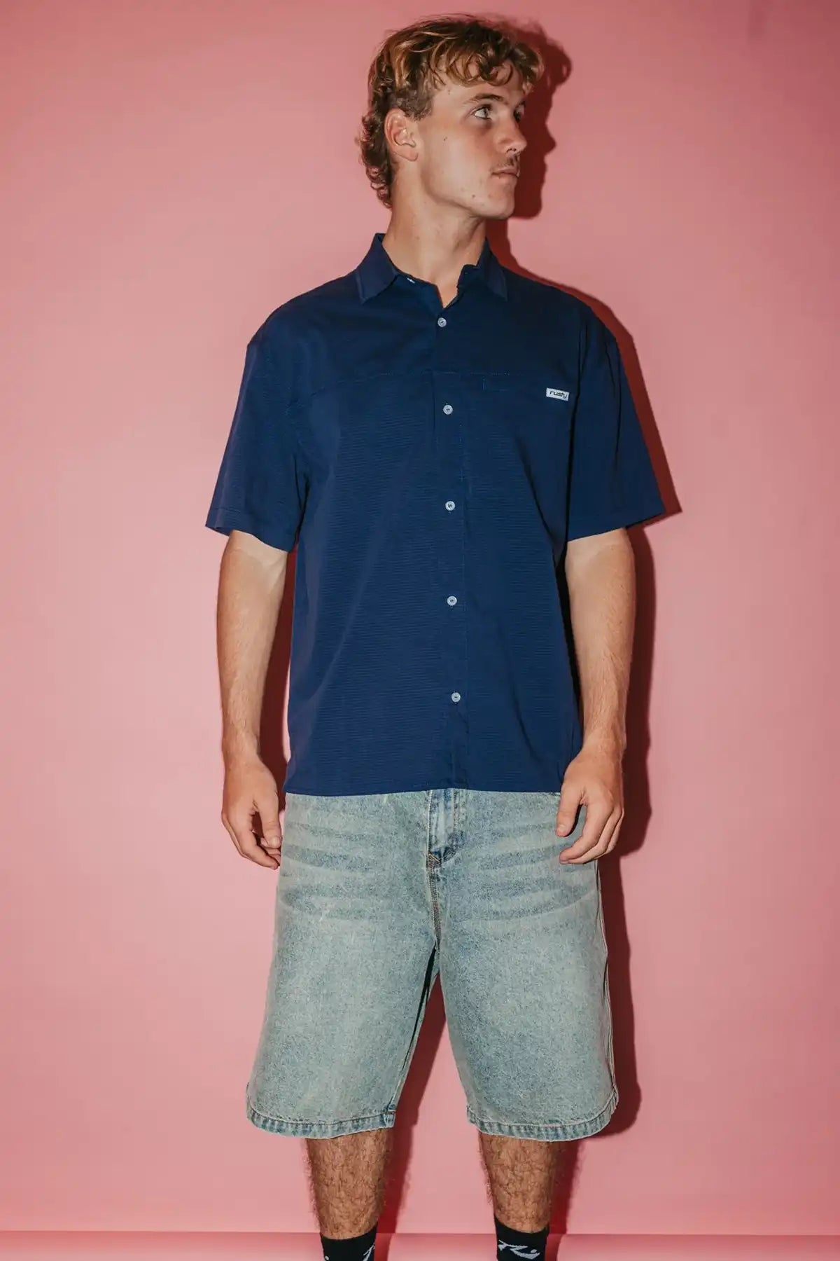 man wearing Navy Blue Rusty Giles Short Sleeve Shirt and denim shorts against a pink background
