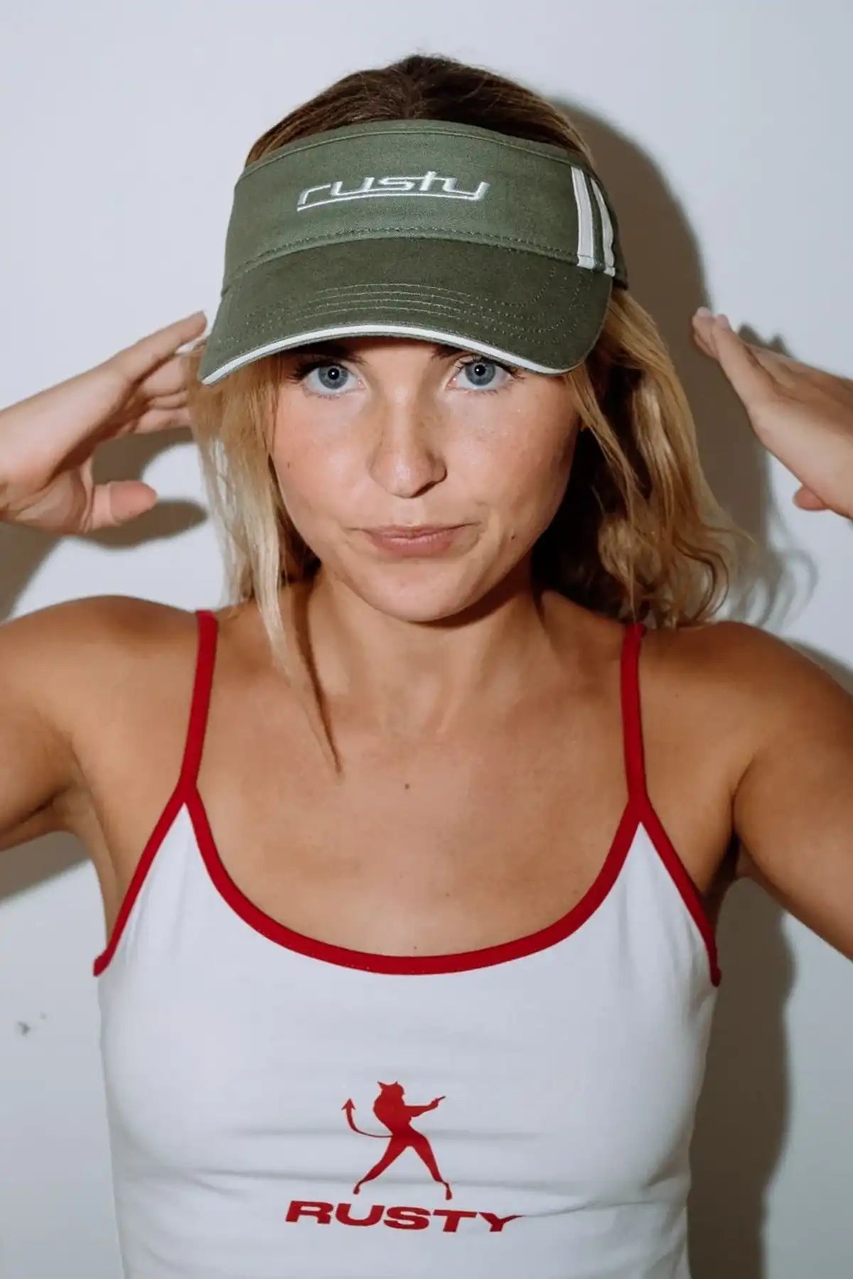 Woman wearing a white tank top with a red logo and a Rusty Love Visor in Arm Green with 'Rusty' branding.