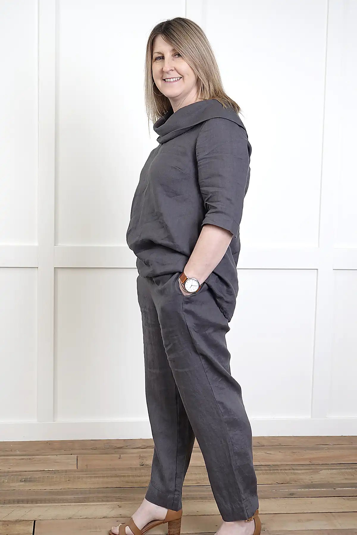 Woman wearing a See Saw Linen 7/8 Flat Front Pant and cowl neck top in Charcoal standing side on against a white paneled wall.