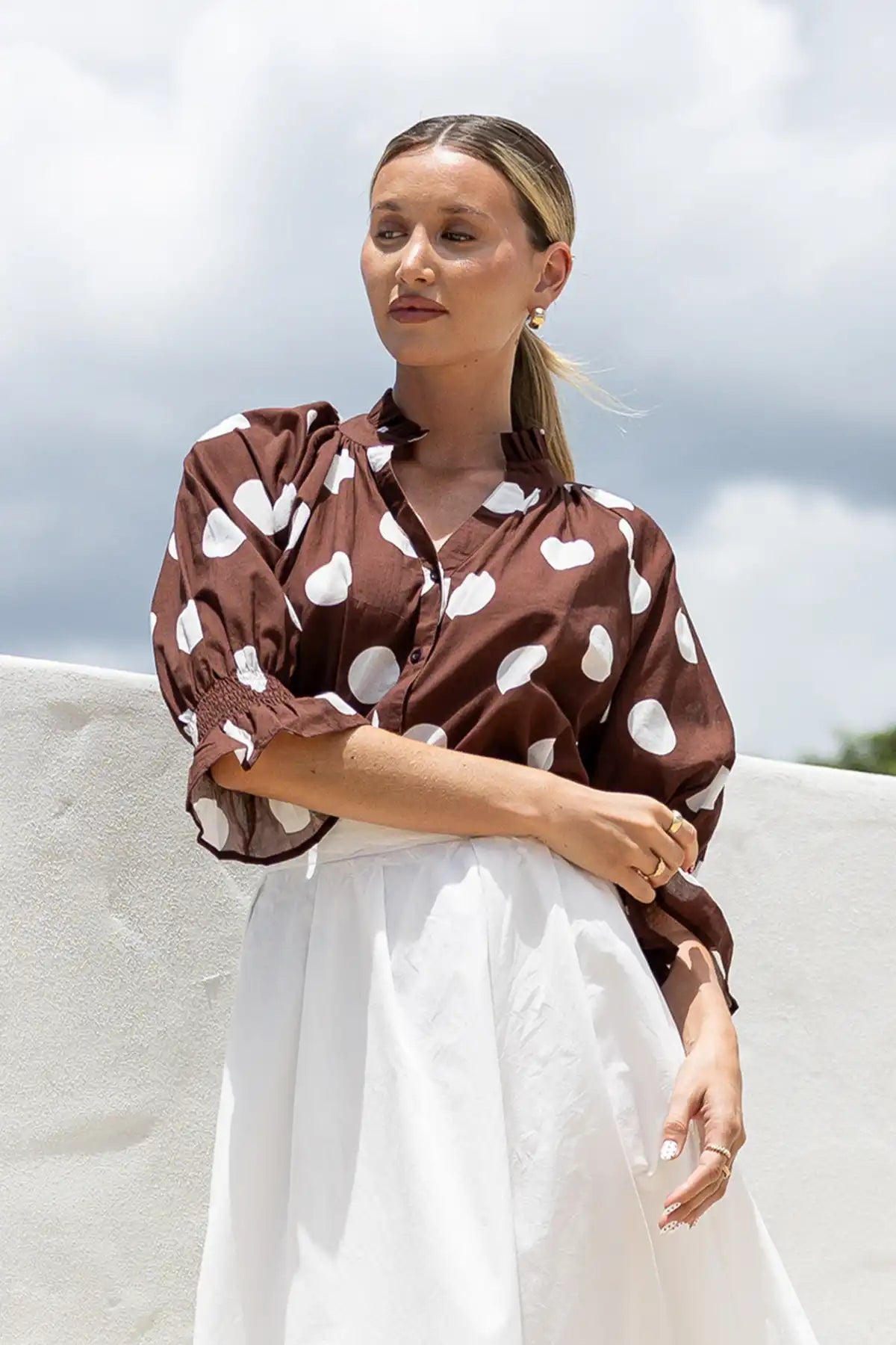 Woman wearing a brown blouse with white heart patterns and a white skirt outdoors.