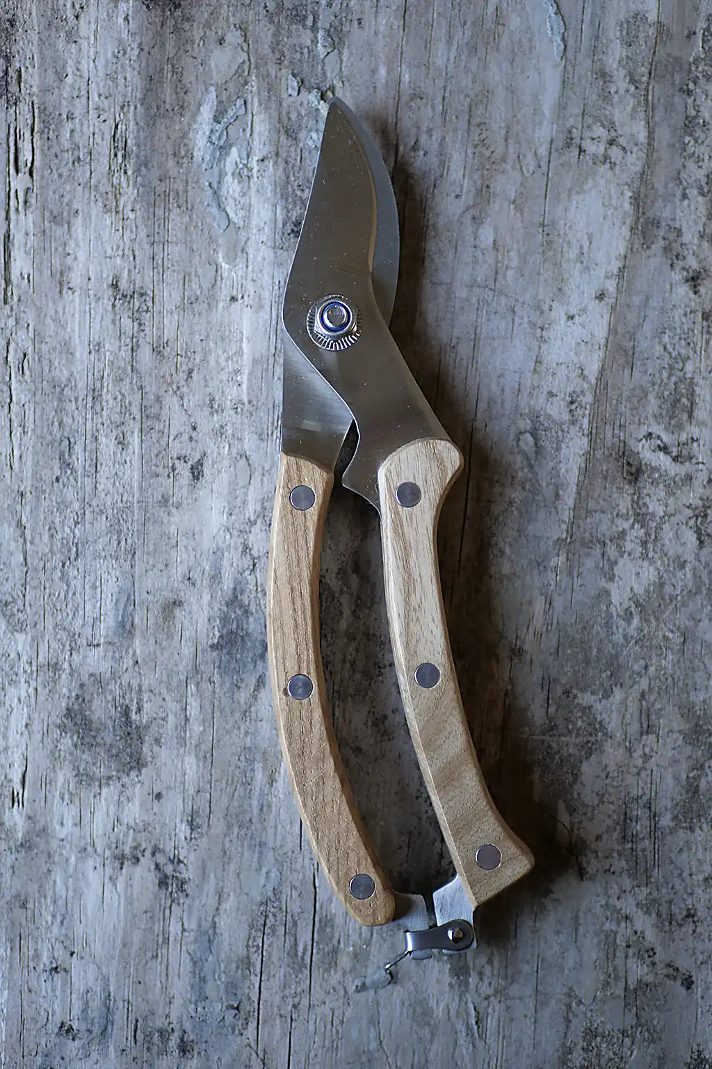 A pair of pruning shears with light-coloured wooden handles and a stainless steel blade, resting on a weathered grey wooden surface.