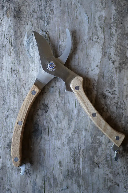 A pair of pruning shears with light-coloured wooden handles and a stainless steel blade, resting on a weathered grey wooden surface.