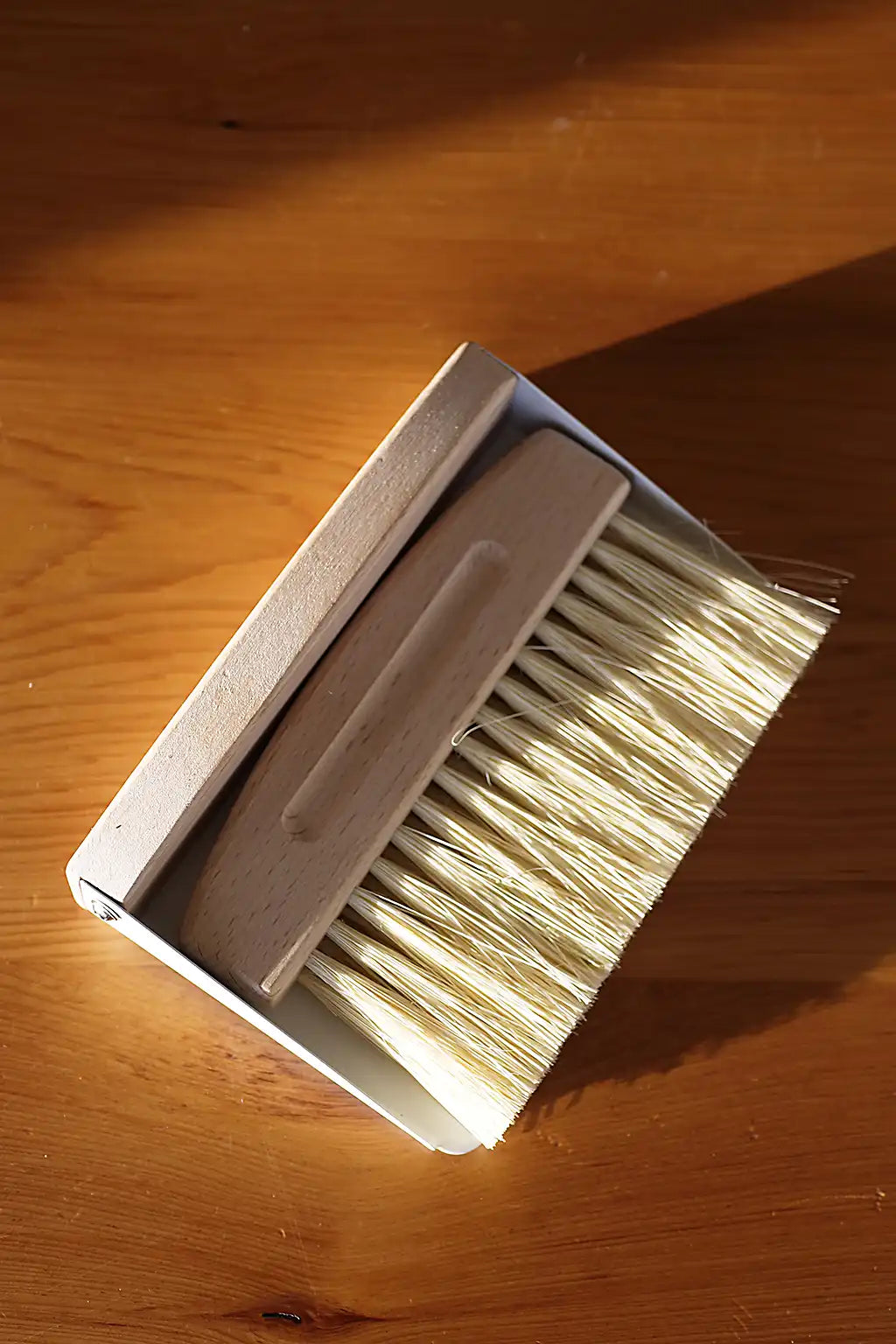 A small cleaning brush with a light wood handle and off-white bristles, photographed on a warm wooden surface in direct sunlight.
