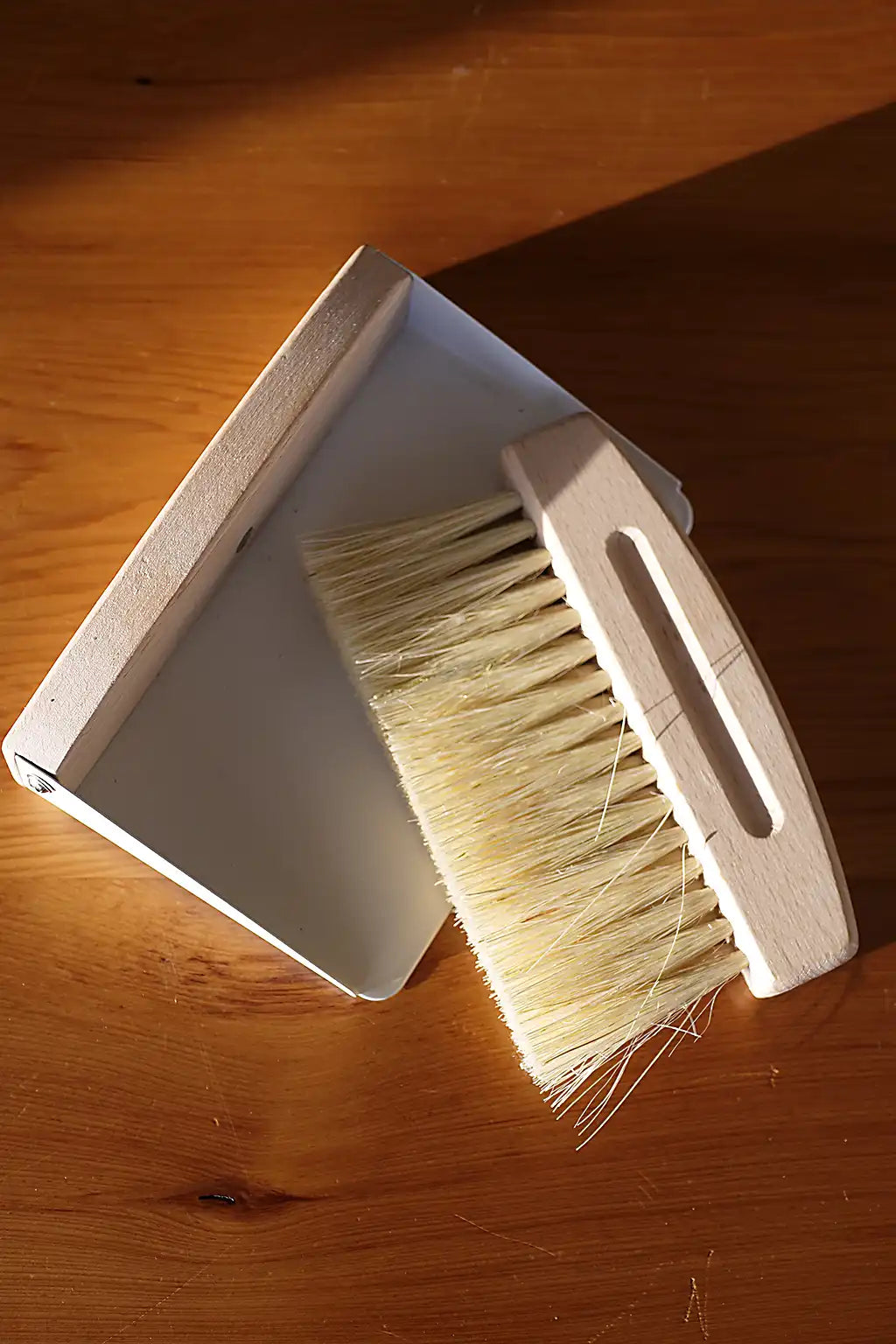 A small wooden cleaning brush with cream-colored bristles and a matching rectangular dustpan, placed on a warm brown wooden surface in direct sunlight.