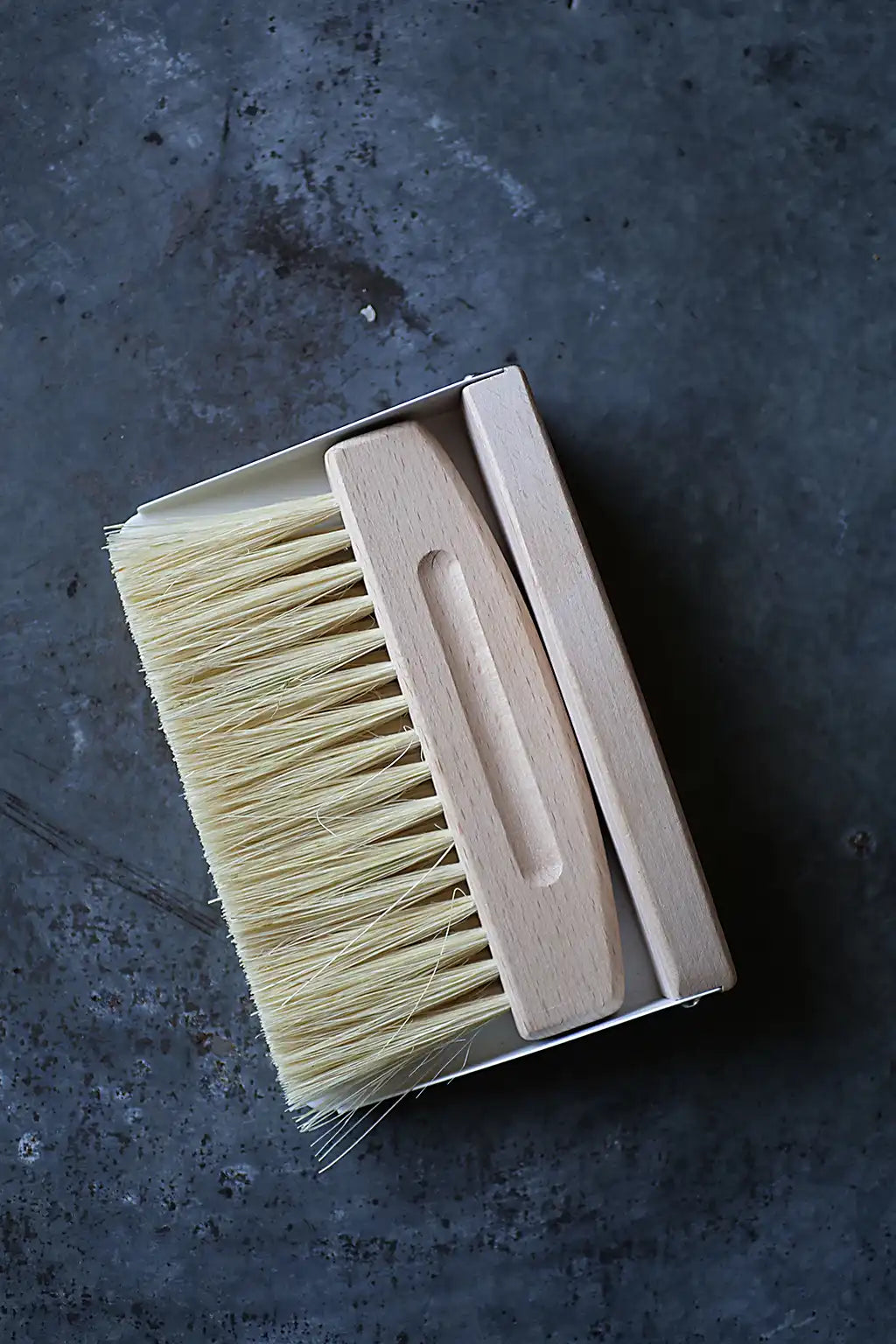 A wooden-handled cleaning brush with pale natural bristles, shown partially out of its open cardboard packaging on a dark textured surface.