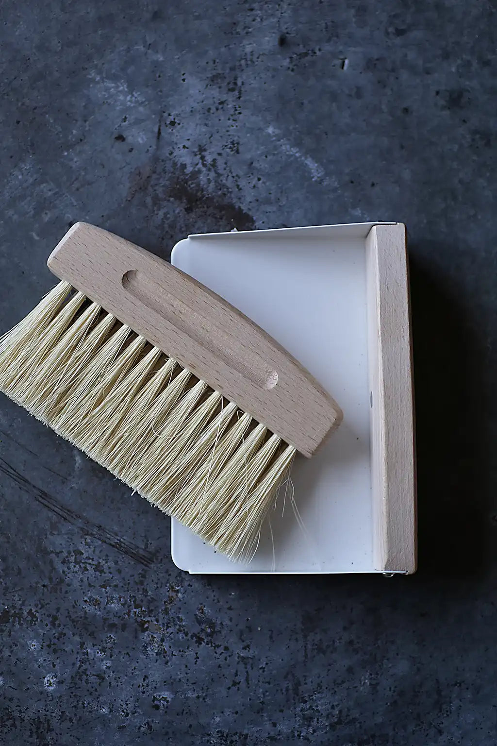 A wooden-handled cleaning brush with natural beige bristles resting on top of a white rectangular box, photographed from above on a dark textured surface.