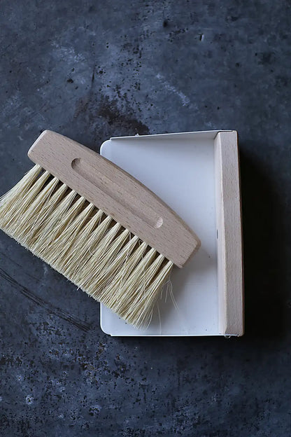 A wooden-handled cleaning brush with natural beige bristles resting on top of a white rectangular box, photographed from above on a dark textured surface.