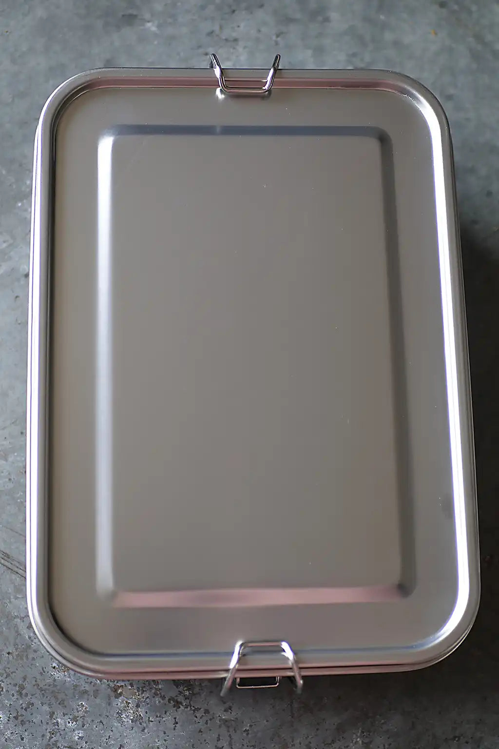 A rectangular stainless steel lidded container  with two metal clasp handles, shown from a top-down perspective on a gray concrete floor.