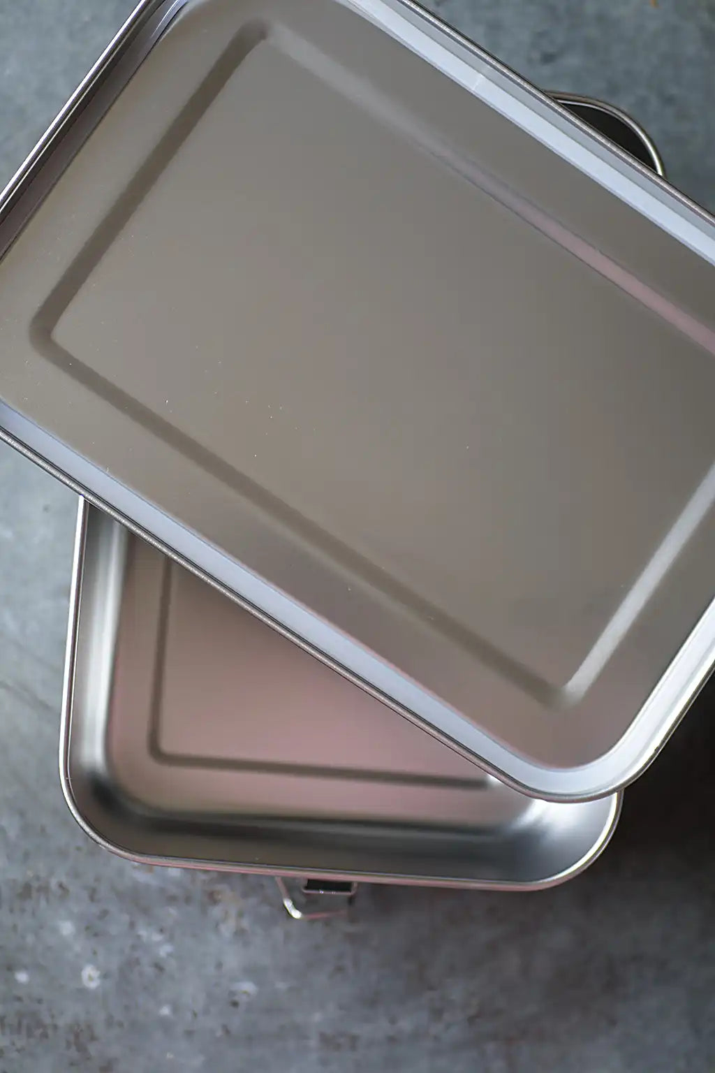 A rectangular metal storage container with lidd, stacked diagonally on a gray textured surface.