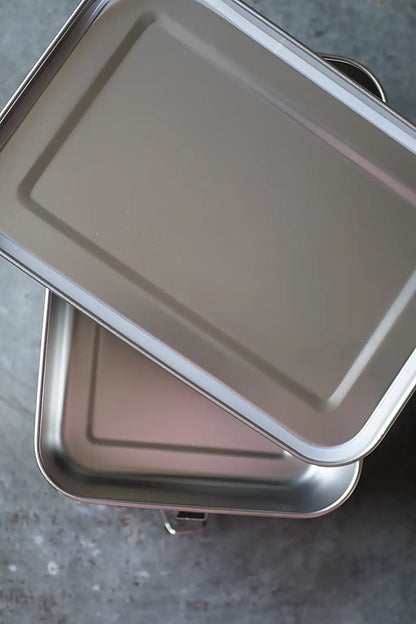 A rectangular metal storage container with lidd, stacked diagonally on a gray textured surface.