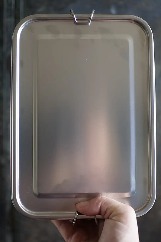 A stainless steel rectangular container with rounded corners and a small locking mechanism at the top and bottom, held by a hand against a dark textured background.