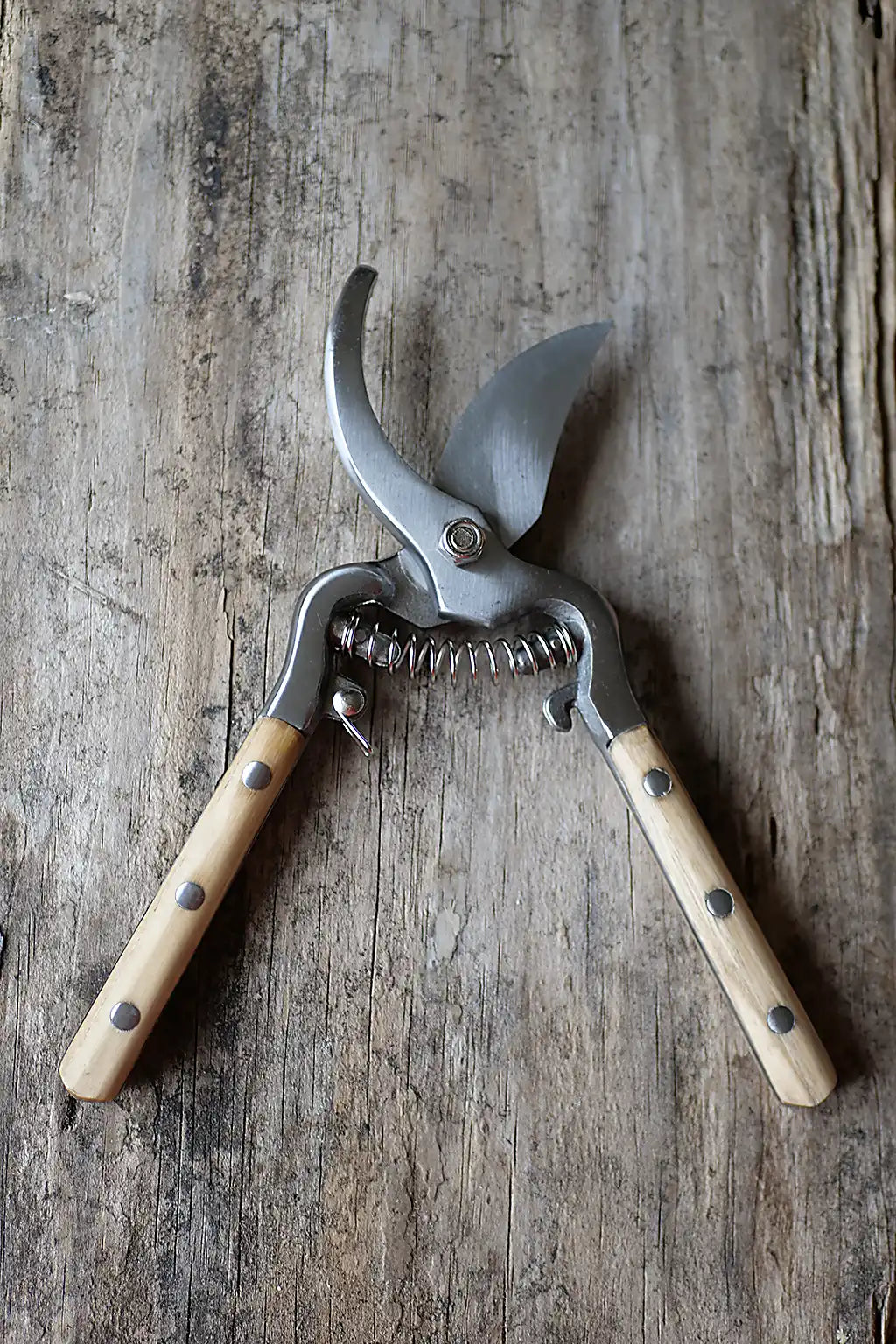 A pair of pruning shears with light wood handles and silver metal blades, resting on a rustic weathered wood surface.