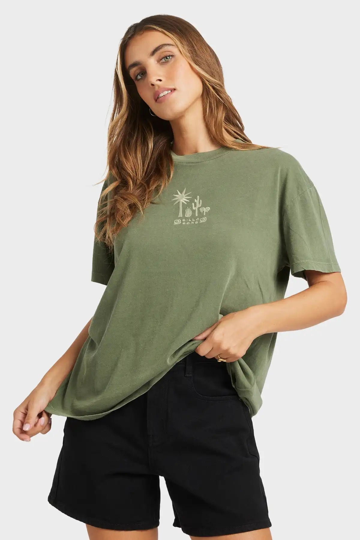 Woman wearing a green Billabong t-shirt with a logo and black shorts on a white background