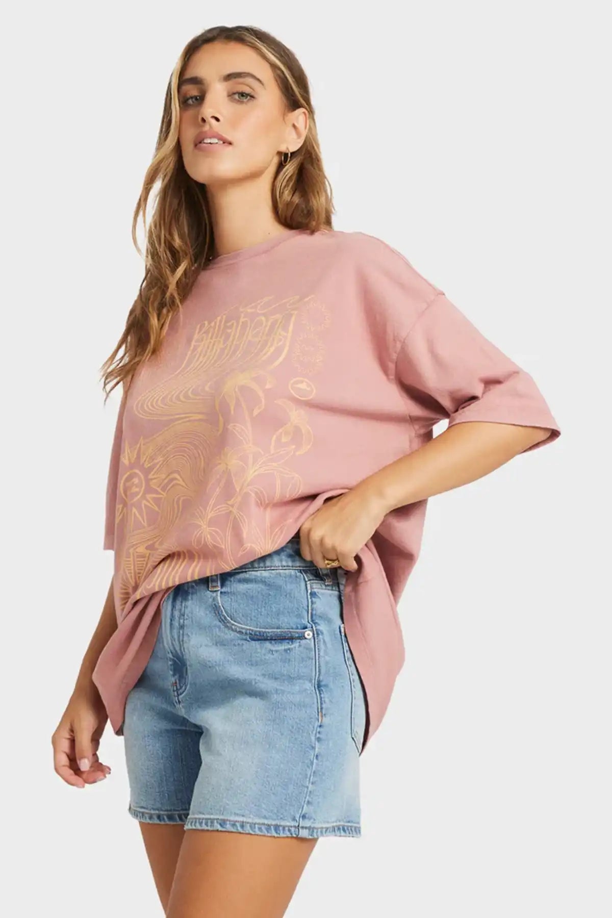 Woman wearing a pink oversized Billabong sweater with gold embroidery and denim shorts on a white background