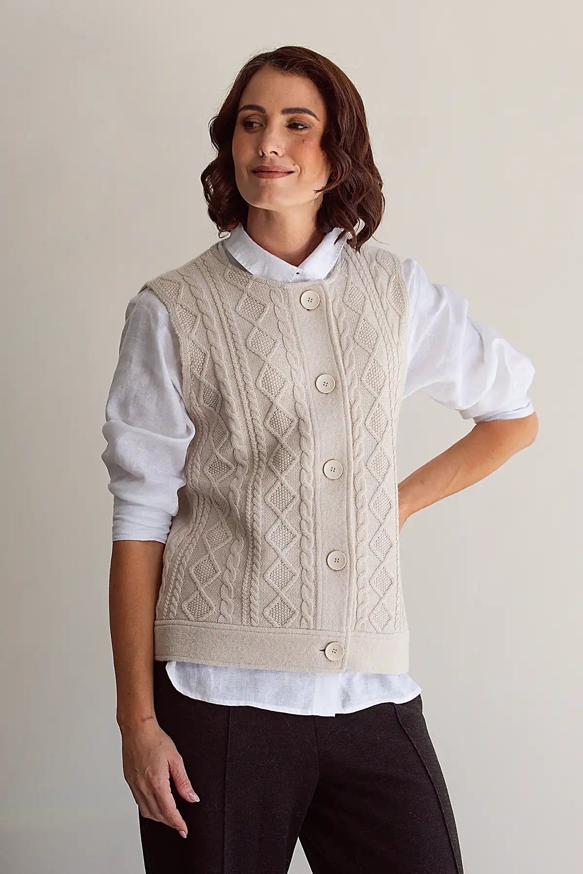 Woman wearing a cream knitted woolen vest over a white shirt with a plain background
