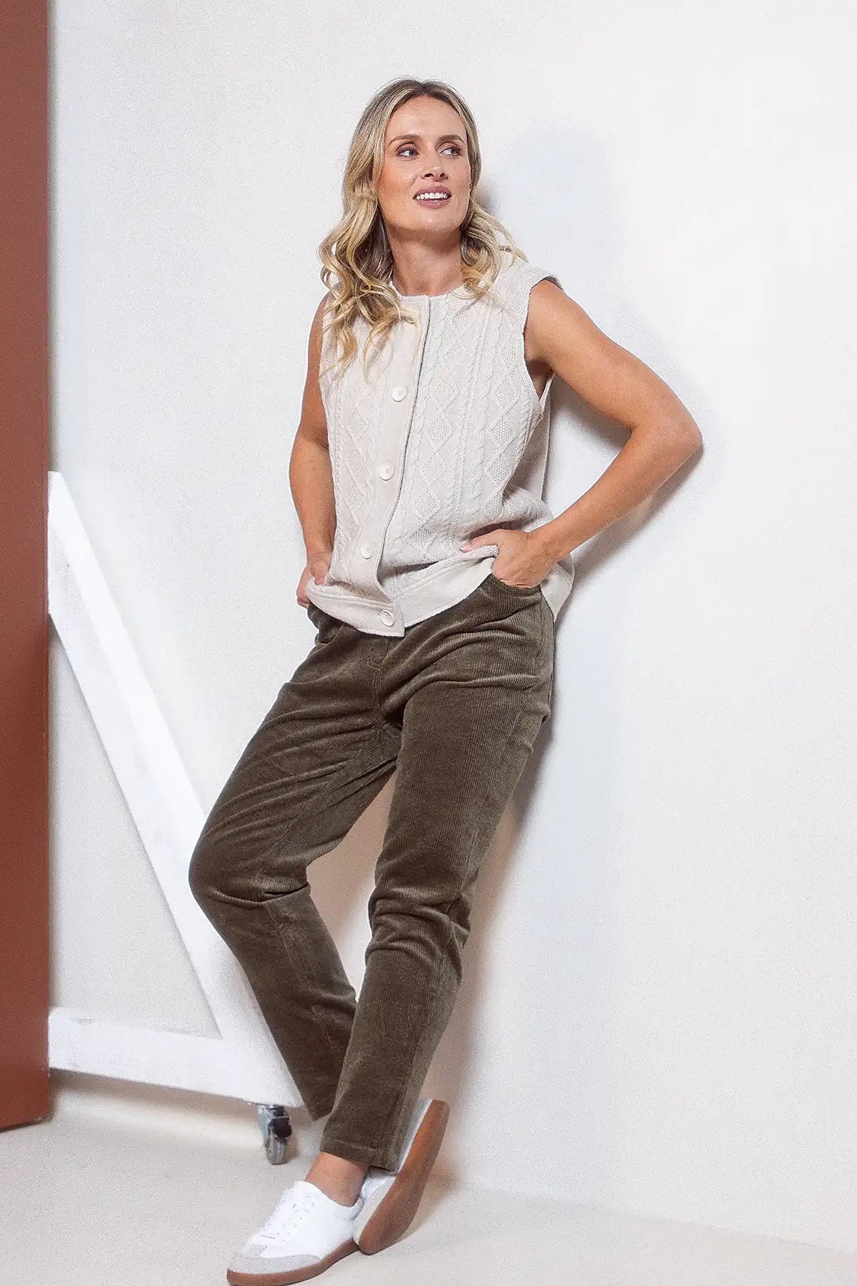 Woman wearing a sleeveless knitted top and brown pants against a white wall.