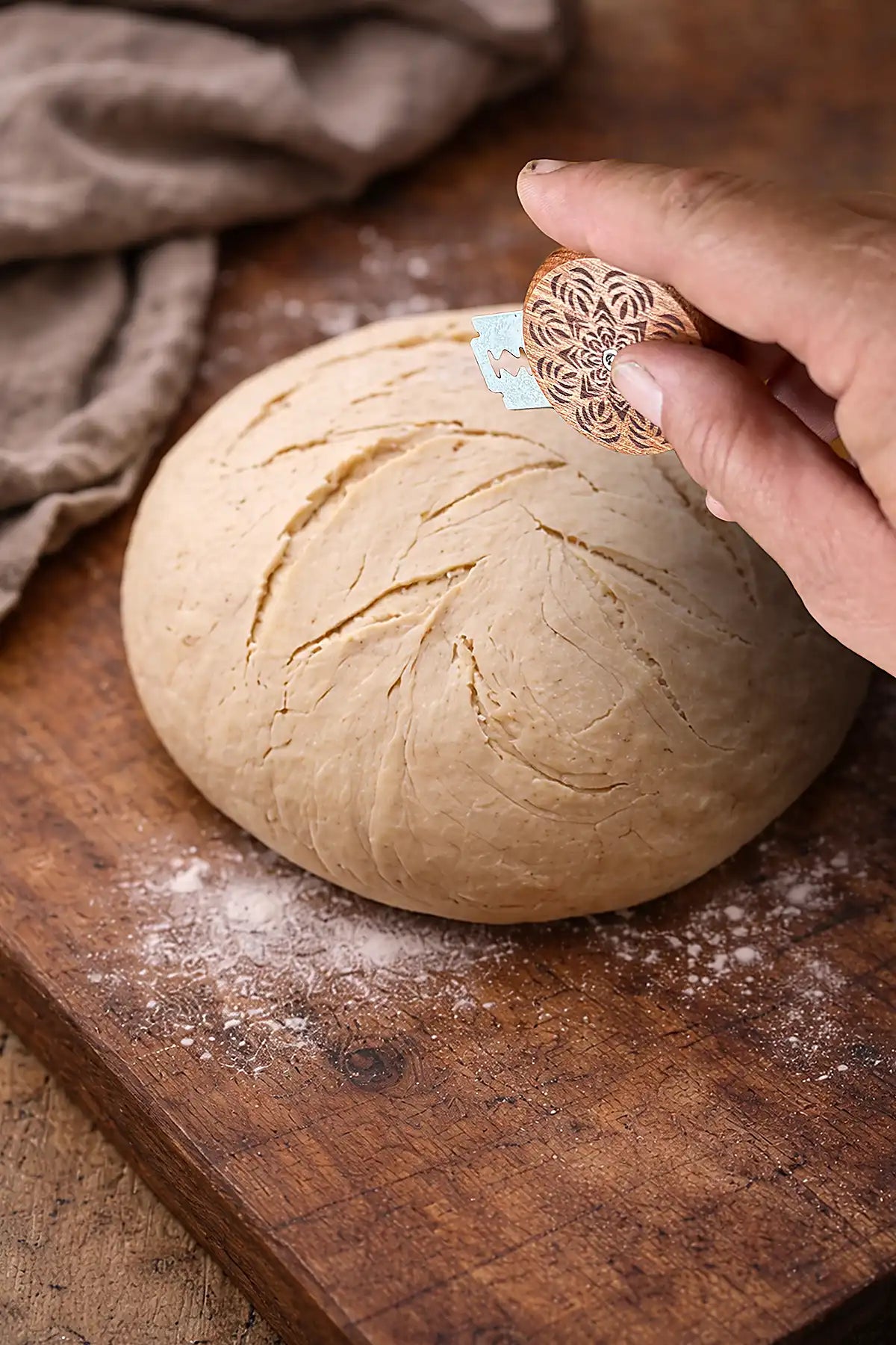 Hand using a wooden bread lame on a ball of dough on a wooden surface.