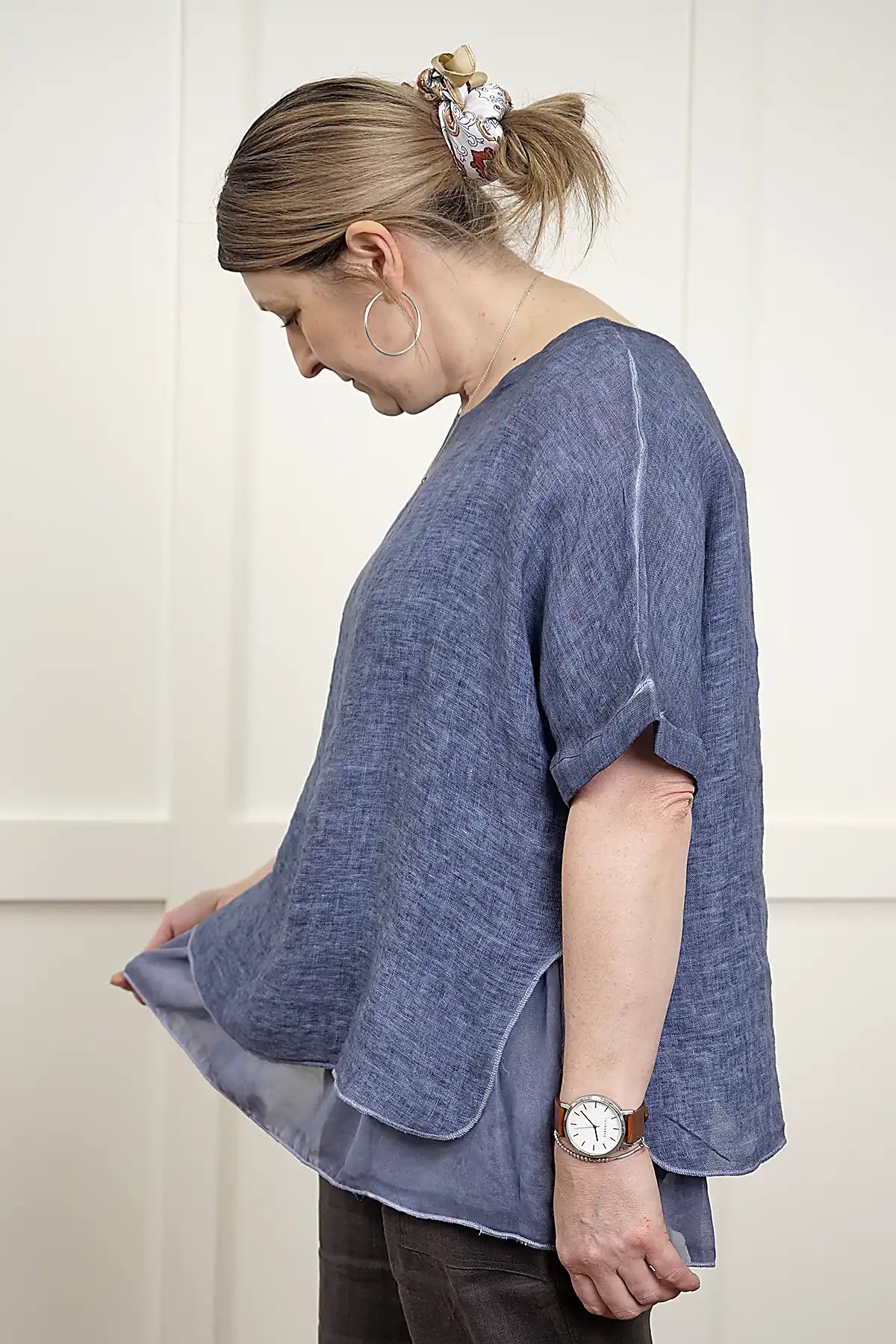 Woman wearing a Elena Linen Top in Denim standing side on looking at the hem against a white background
