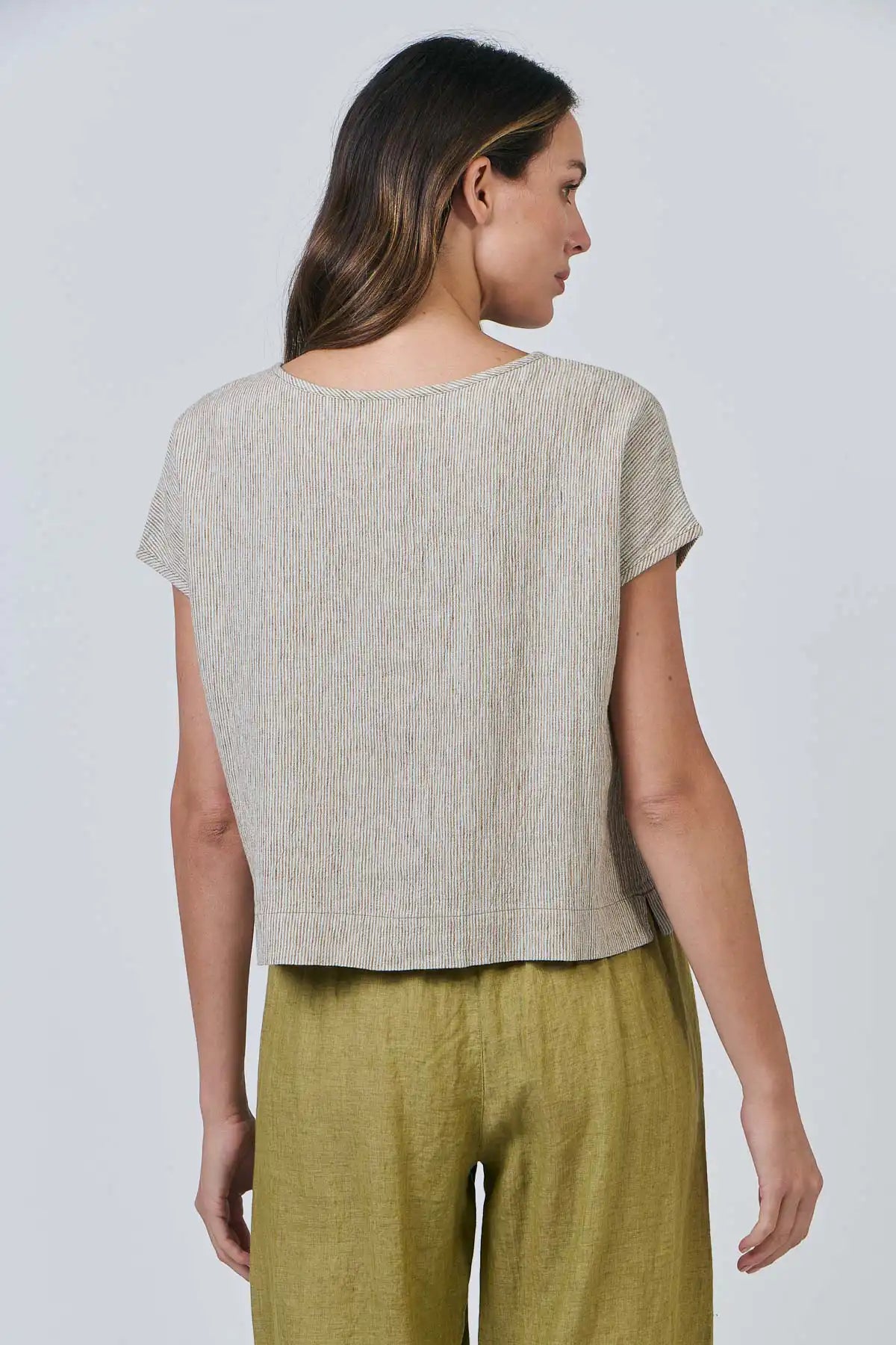 Woman wearing a Elysian Linen Top – Blister Stripe and green pants against a white background rear view