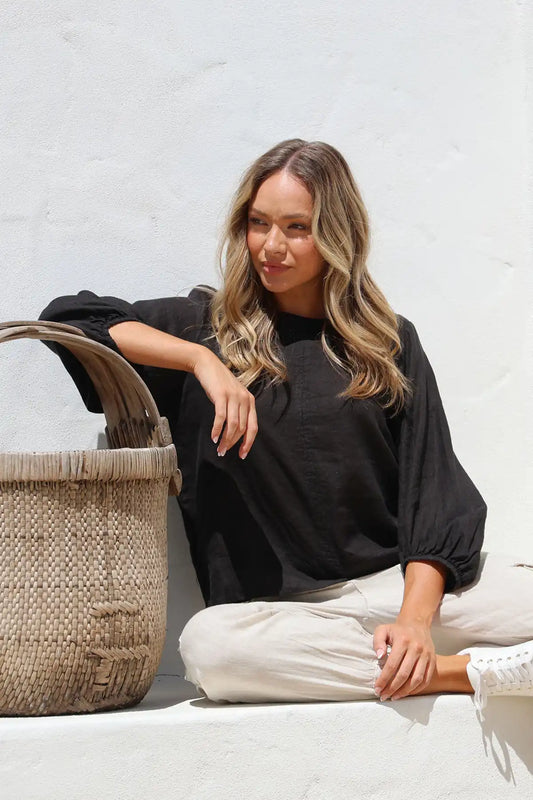 Woman sitting on a white surface with a woven basket next to her against a light gray wall wearing the Giovanna Italian Linen Top in black