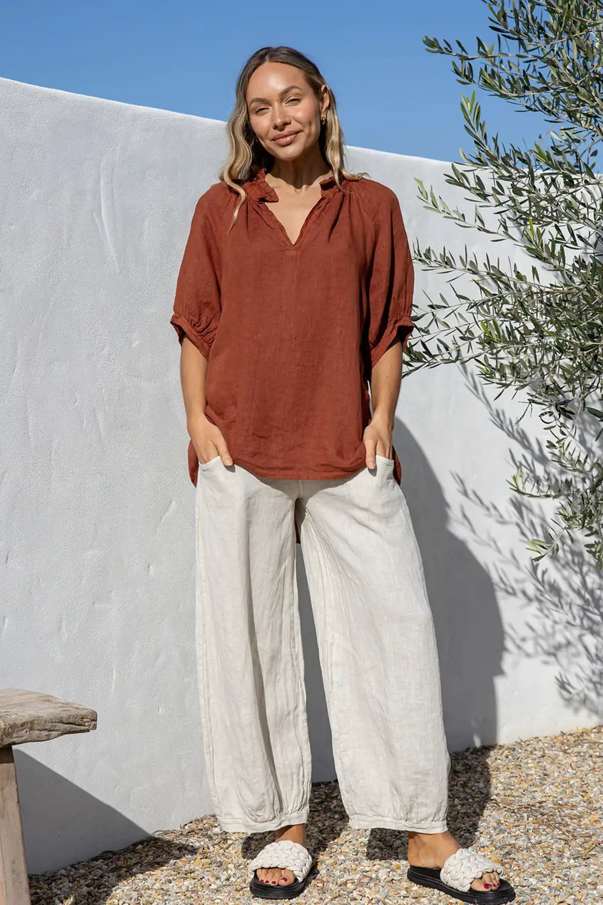 Woman wearing a Giulia Italian Linen Coco Top and white pants standing against a white wall with greenery.