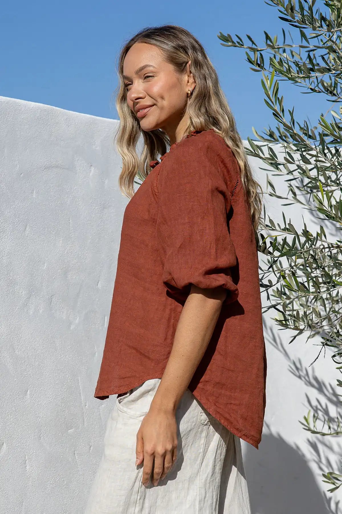 Woman wearing a Giulia Italian Linen Coco Top and natural pants standing side on against a white wall with greenery.