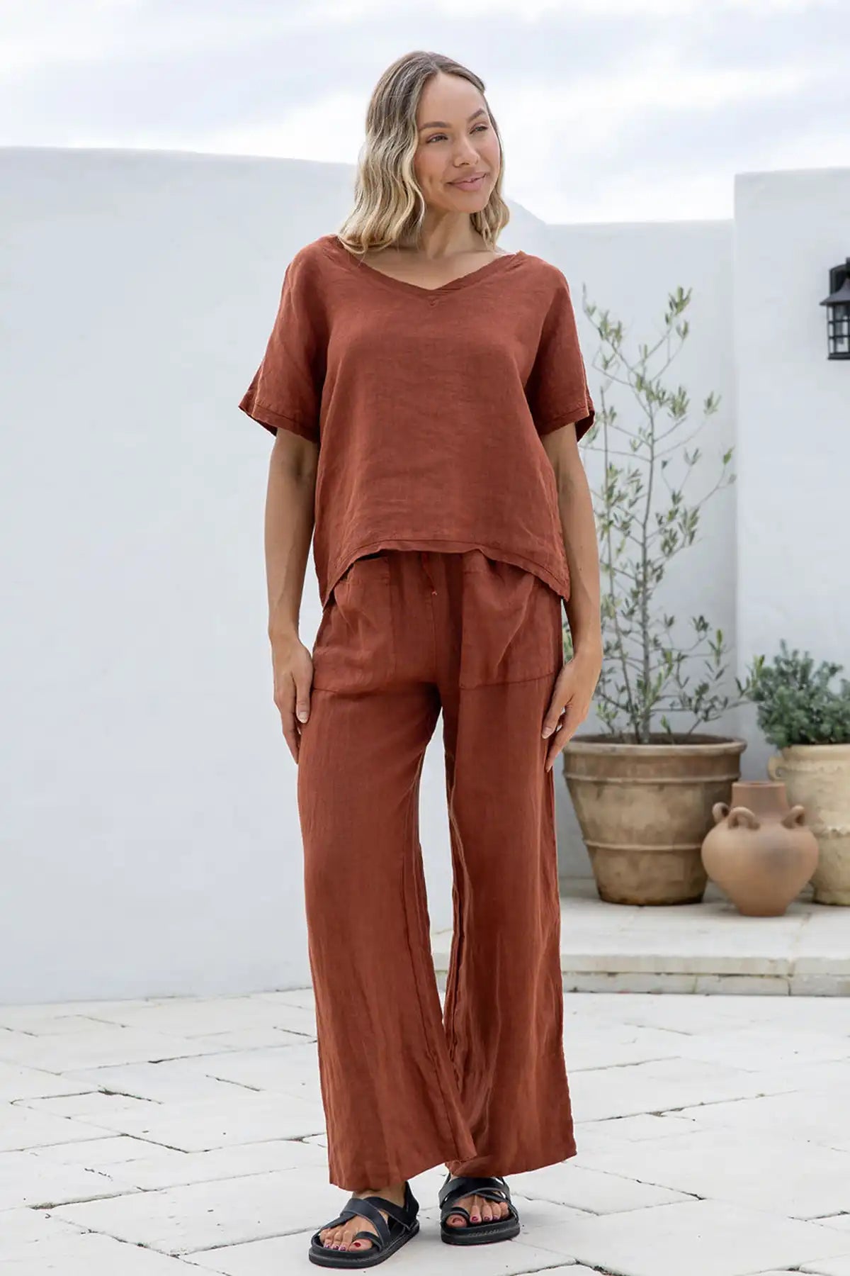Woman wearing a Lucia Italian Linen Coco Top and coco pants standing outdoors with white walls and potted plants in the background.