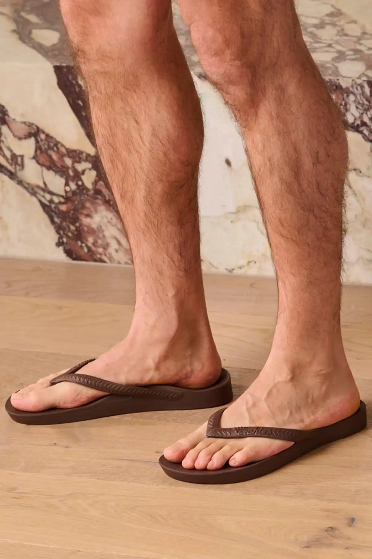 Person wearing brown sandals on a wooden floor with a patterned wall in the background