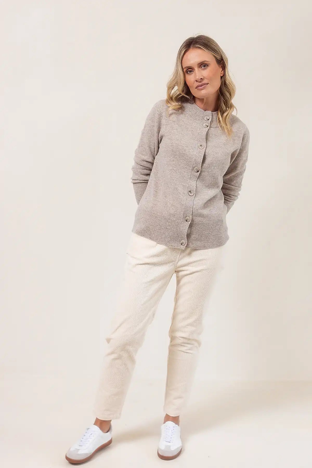 Woman wearing a wheat cardigan, white pants, and white shoes on a beige background