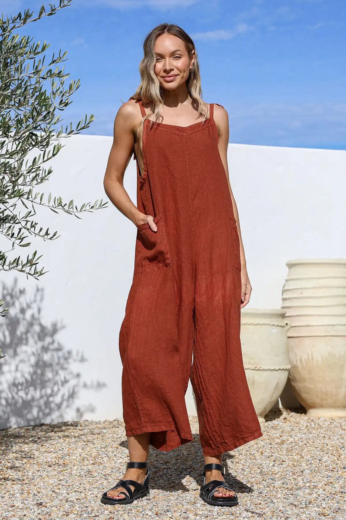 Woman wearing a Mia Italian Linen Coco Wide Leg Jumpsuit standing outdoors with a white wall and potted plant in the background.