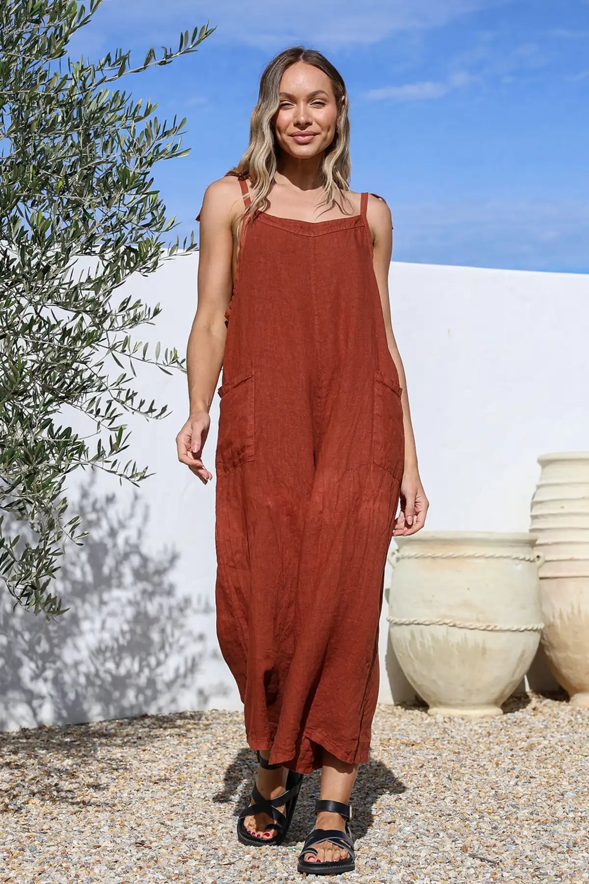 Woman wearing a Mia Italian Linen Coco Wide Leg Jumpsuit standing outdoors with a white wall and olive tree in the background.
