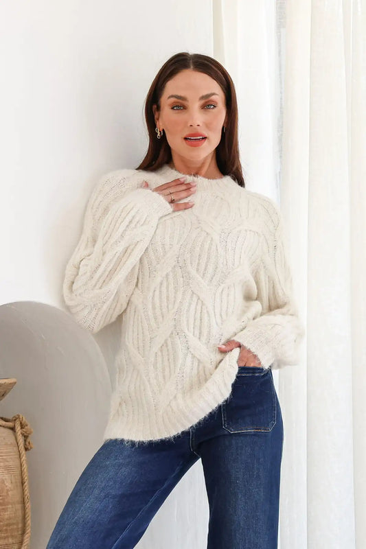 Woman wearing a white cable knit sweater and blue jeans indoors.