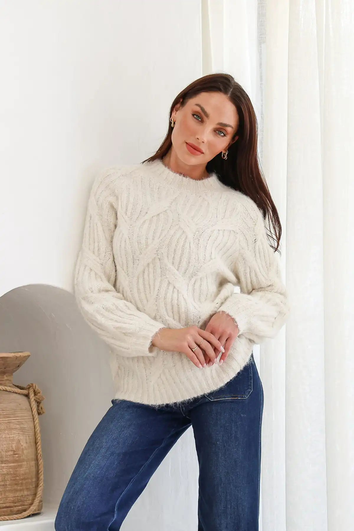 Woman wearing a cream sweater and blue jeans standing against a white wall.