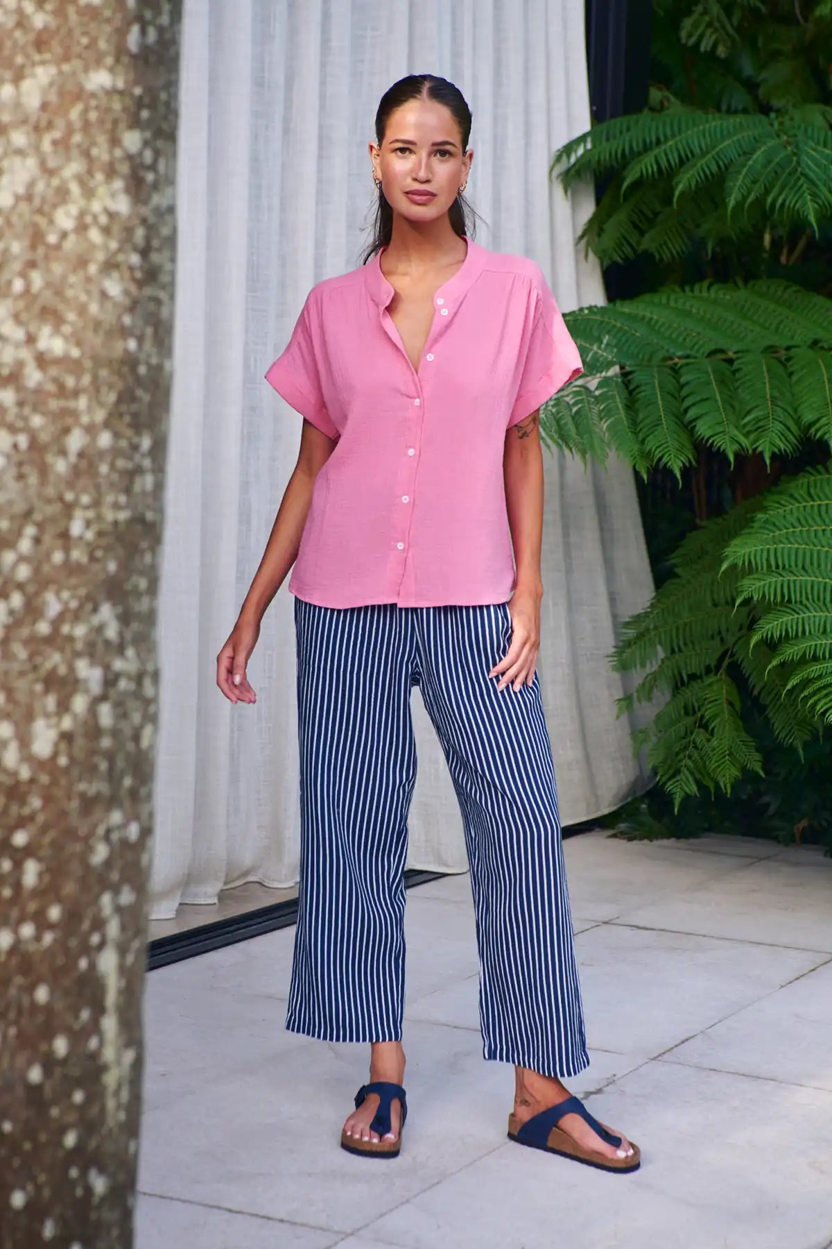 Woman wearing a pink shirt and blue striped pants standing outdoors with plants in the background