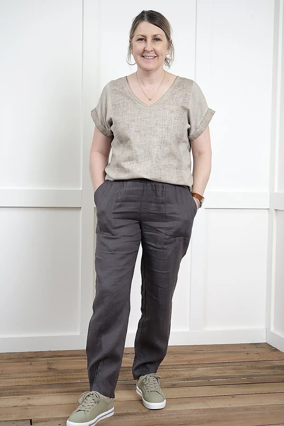 Woman wearing a Rosa V Neck Linen Top in Latte and dark pants standing in a room with white walls and wooden floor.