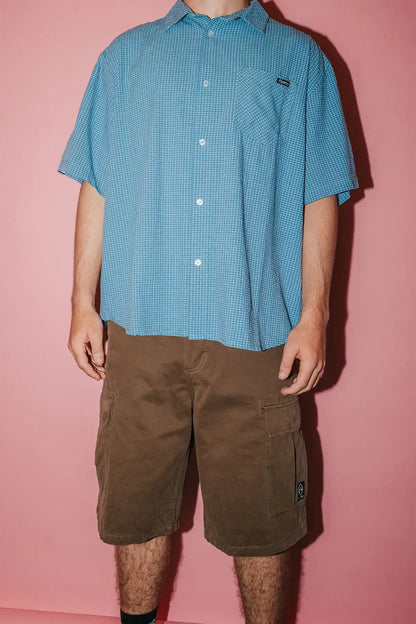 Person wearing a blue checkered shirt and Rusty Commando 23" Baggy Cargo Short in Pinecone against a pink background