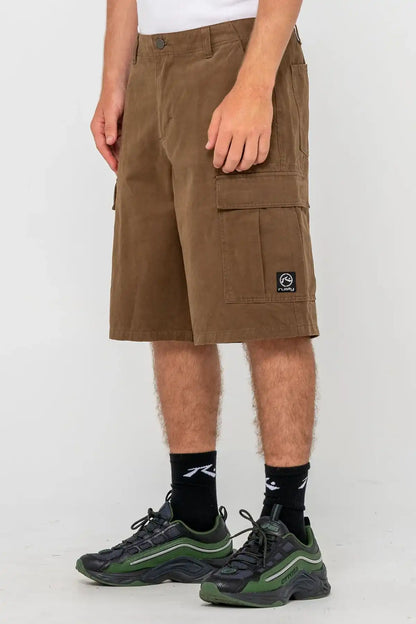 Person wearing Rusty Commando 23" Baggy Cargo Short in Pinecone, side view with a visible brand logo on a white background
