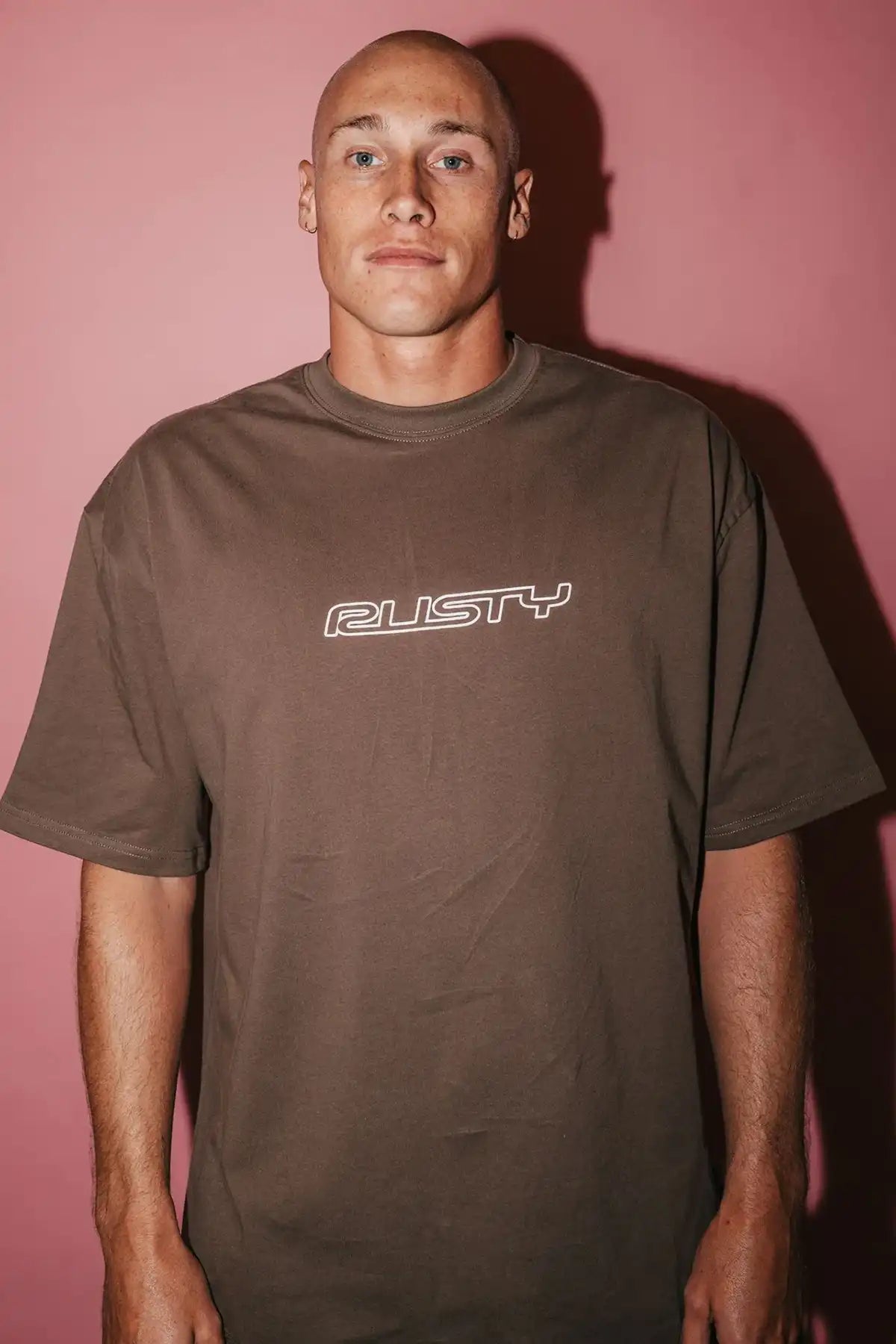 Man wearing a brown t-shirt with 'Rusty' logo against a pink background