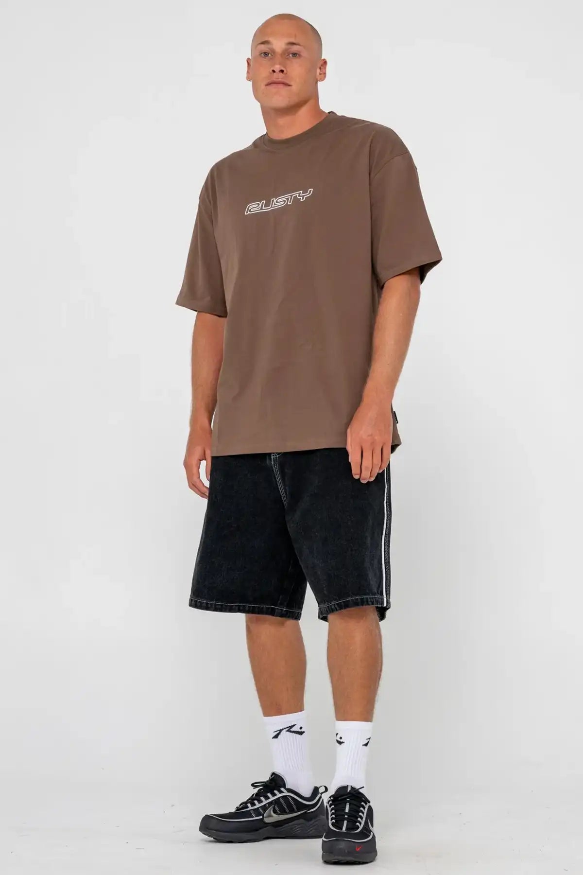 Man wearing a brown Rusty t-shirt with text, black shorts, and white socks on a white background