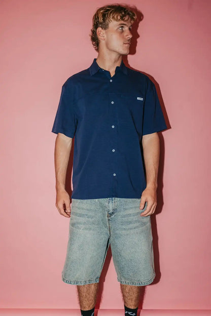 man wearing Navy Blue Rusty Giles Short Sleeve Shirt and denim shorts against a pink background
