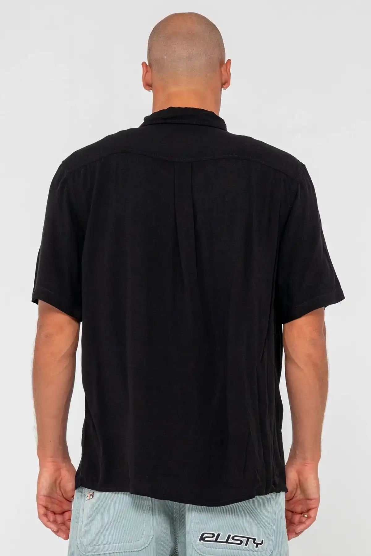 man wearing a black Rusty Mens Razor Blade Rayon Shirt and denim shorts on a pink background standing back on